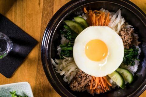 Korean bibimbap served in a black bowl with white rice, sautéed vegetables, minced meat, and a sunny-side-up egg on top, placed on a wooden table.