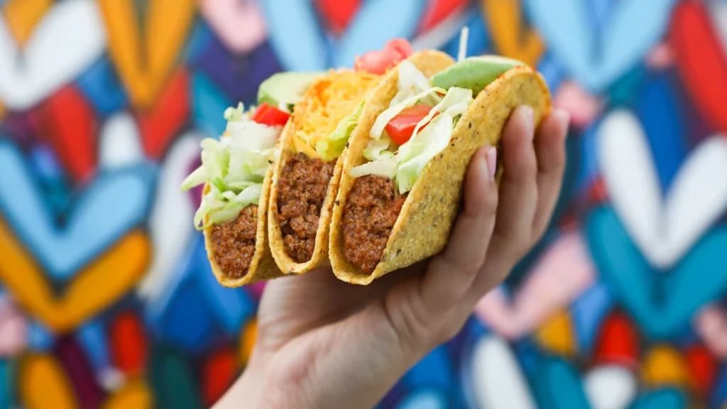 Hand holding three vegetarian tacos with lettuce, tomato, cheese, and plant-based meat, against a colorful graffiti background.