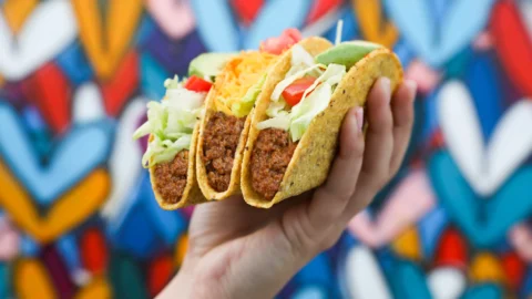Hand holding three vegetarian tacos with lettuce, tomato, cheese, and plant-based meat, against a colorful graffiti background.