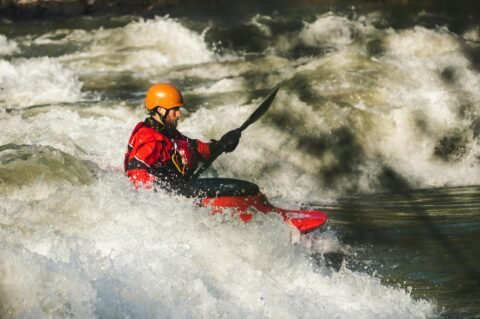 Man kayaking through intense white-water rapids, wearing a red jacket and orange helmet, showcasing an extreme adventure activity in a wild river setting.