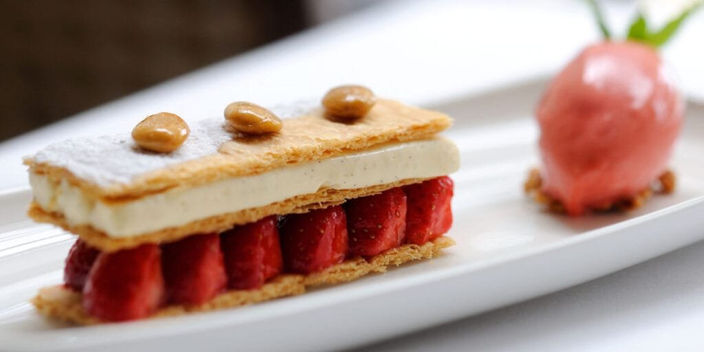Elegant French dessert featuring a mille-feuille layered with puff pastry, vanilla cream, and fresh strawberries, garnished with glazed nuts and served with a scoop of pink sorbet on a white plate.
