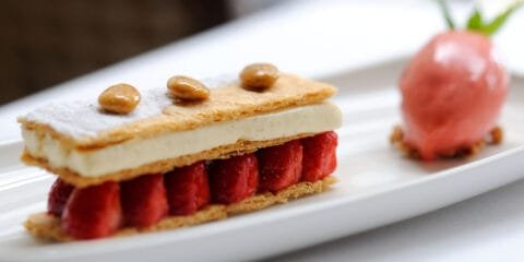 Elegant French dessert featuring a mille-feuille layered with puff pastry, vanilla cream, and fresh strawberries, garnished with glazed nuts and served with a scoop of pink sorbet on a white plate.
