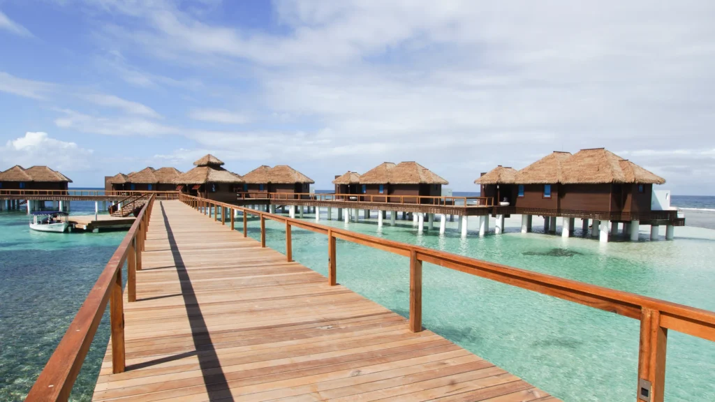 Scenic view of Palafitos Overwater Bungalows in Mexico with thatched roofs, clear turquoise water, and a wooden walkway leading over the ocean under a sunny sky.