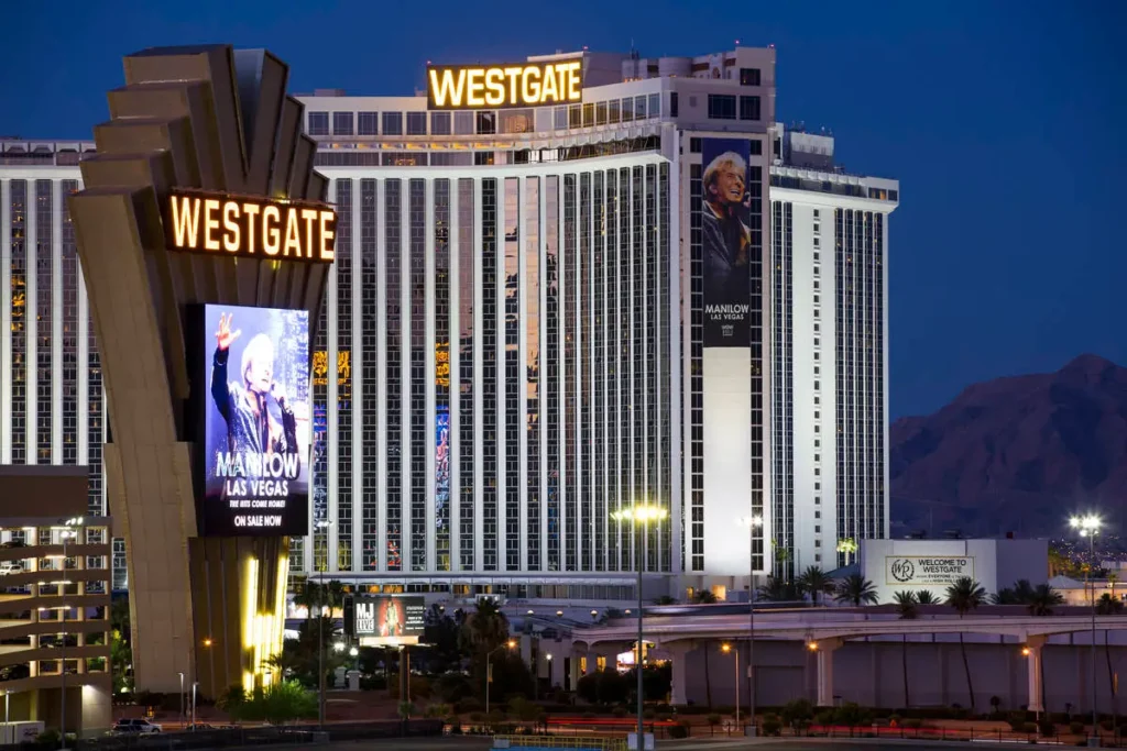 The illuminated facade of Westgate Las Vegas Resort and Casino at night, featuring tall white towers, glowing signage, and a large digital screen of Barry Manilow.