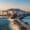 Coastal view of Naxos Town, Greece, with whitewashed buildings on a hill, a stone pathway along the sea, and waves crashing against the rocky shoreline at sunset.