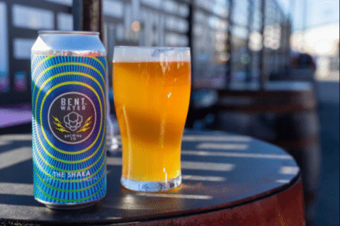 Bent Water Brewing Co. beer can and pint glass of golden ale on an outdoor table.