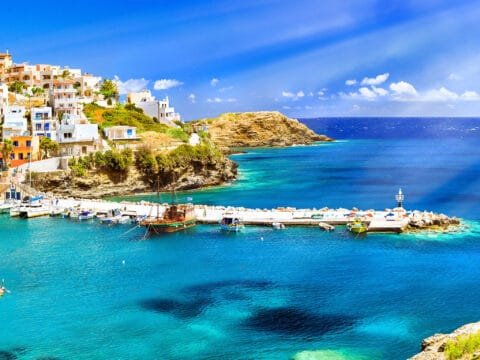 Vibrant coastal view of a harbor in Crete, Greece, featuring turquoise waters, anchored boats, a traditional wooden ship, and colorful hillside houses under a bright blue sky.
