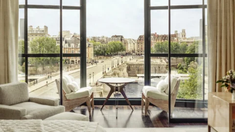Elegant hotel room interior with two lounge chairs and a table facing large floor-to-ceiling windows, offering a panoramic view of a historic Parisian street, bridge, and classic buildings along the Seine River.