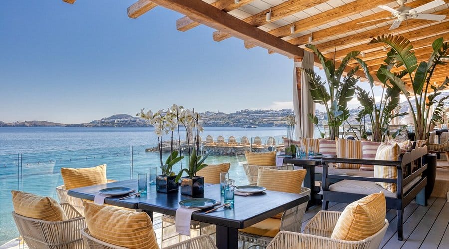 Elegant beachfront restaurant at Buddha-Bar Mykonos with tropical decor, cushioned seating, and panoramic views of the Aegean Sea under a wooden pergola.