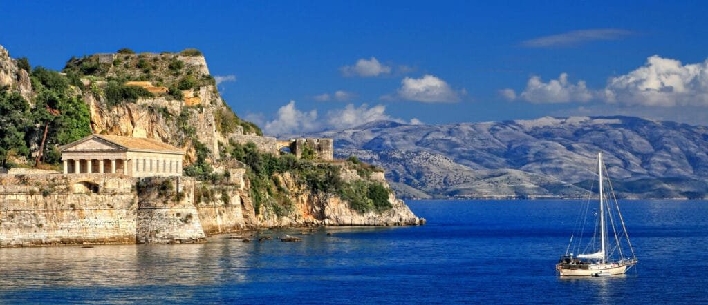 Discover Corfu Greece - An Island of Dreamy Beaches, Seaside Villages, Flamingos & Five-Star Escapes 4 View of a sailboat floating near the Old Fortress of Corfu, with a neoclassical temple structure built into the rocky cliffs and mountains in the background under a clear blue sky.