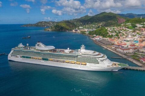 Large Royal Caribbean cruise ship "Jewel of the Seas" docked at a scenic Mediterranean port, surrounded by turquoise waters and a colorful coastal town with lush green hills.
