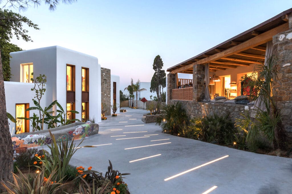Stylish evening view of Nammos Village in Mykonos, featuring whitewashed Cycladic buildings, modern boutiques, warm lighting, and landscaped walkways with mosaic accents.