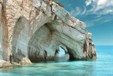 9 Cheapest Places in Greece to Go on Holiday For Budget Travelers 92 Natural limestone sea arch formations over clear turquoise waters along the coast of Zakynthos, Greece, under a bright blue sky with wispy clouds.