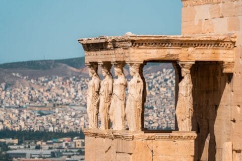 10 Best Places to Visit in Greece for Stories You'll Tell Forever 19 Close-up view of the Caryatids, the sculpted female columns of the Erechtheion temple on the Acropolis of Athens, with the city of Athens and surrounding hills in the background.