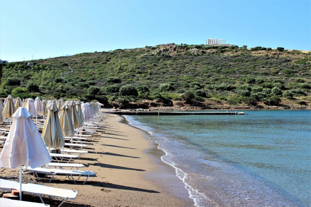 Best 9 Athens Greece Beaches for Athenian Sun & Sea Lovers 4 Organized sandy beach with rows of white sunbeds and umbrellas along calm turquoise waters, set against a lush green hillside with the Temple of Poseidon visible in the distance at Cape Sounion, Athens, Greece.
