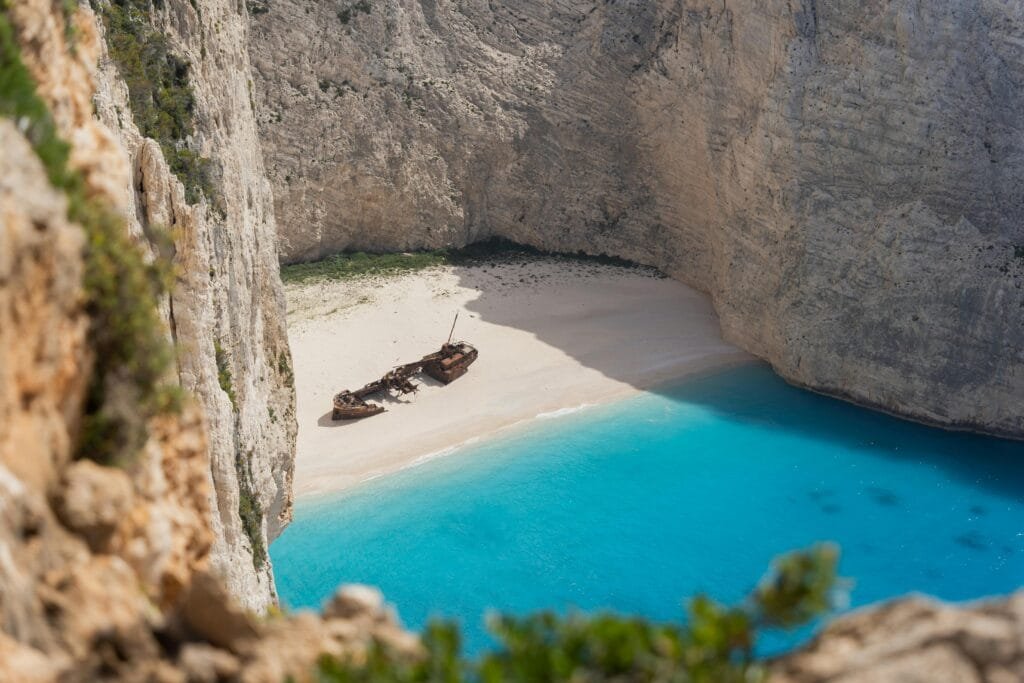 Aerial view of Navagio Beach (Shipwreck Beach) on Zakynthos, Greece, featuring a rusted shipwreck resting on white sand surrounded by towering limestone cliffs and turquoise-blue water.