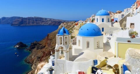 Iconic view of Santorini, Greece, featuring whitewashed buildings with blue-domed churches perched on cliffs overlooking the deep blue Aegean Sea and caldera.