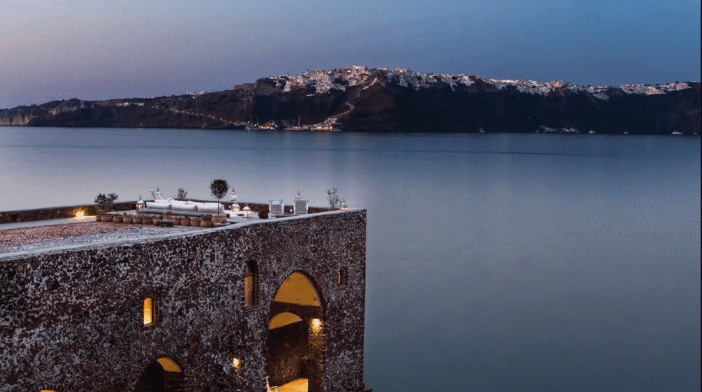 Cliffside stone terrace with cozy seating and lanterns overlooking the calm Aegean Sea at dusk, with the illuminated village of Santorini visible across the water.