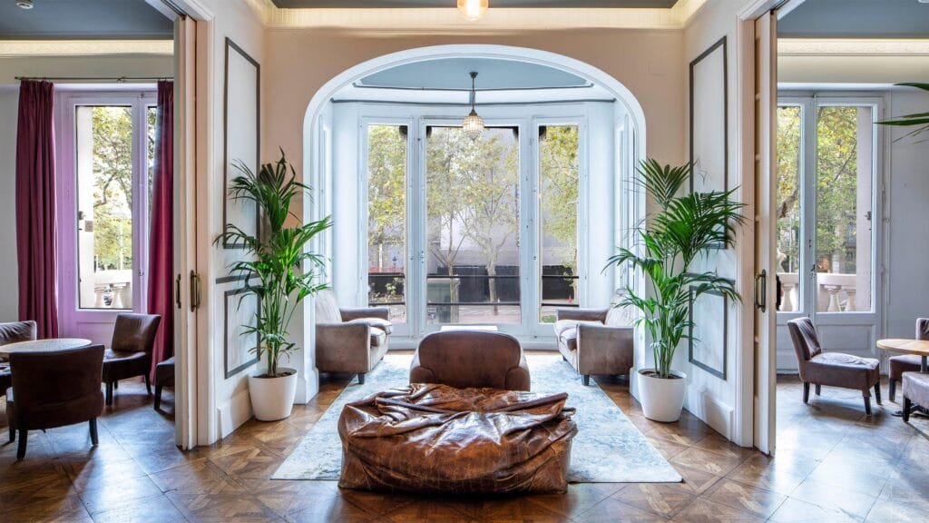 10 Best Youth Hostels in Barcelona for Your 2025 Catalan Adventure 4 Elegant common area in a Barcelona hostel with large windows, vintage wooden floors, indoor plants, and cozy lounge seating including leather chairs and a bean bag.