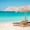 Two wooden sun loungers with straw umbrellas on a sandy beach facing the turquoise waters of Mykonos, Greece, with a rocky hill in the background under a clear blue sky.