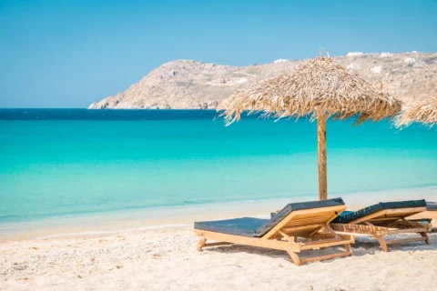 Two wooden sun loungers with straw umbrellas on a sandy beach facing the turquoise waters of Mykonos, Greece, with a rocky hill in the background under a clear blue sky.